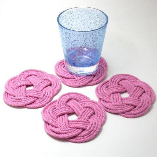 A blue glass rests on one of four true Nautical Sailor Knot Coasters Set of 4 in pink, all handmade and neatly arranged on a white surface. by Sea Bags