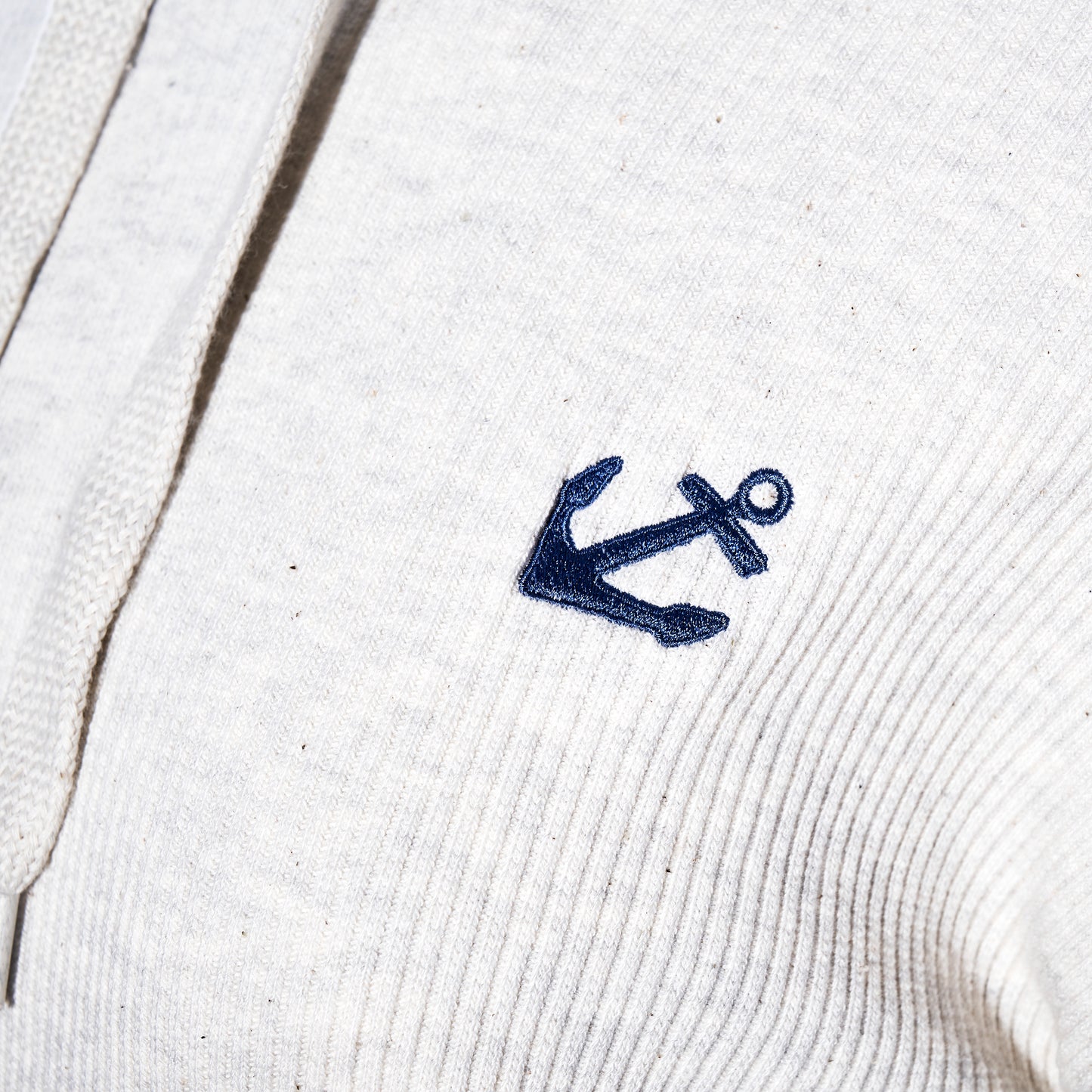 Close-up of the Soft As A Grape Ribbed Hoodie Pullover - Oatmeal Anchor, featuring a light-colored ribbed design and a navy blue embroidered anchor on the chest, perfect for adding a touch of coastal style to your look. by Sea Bags