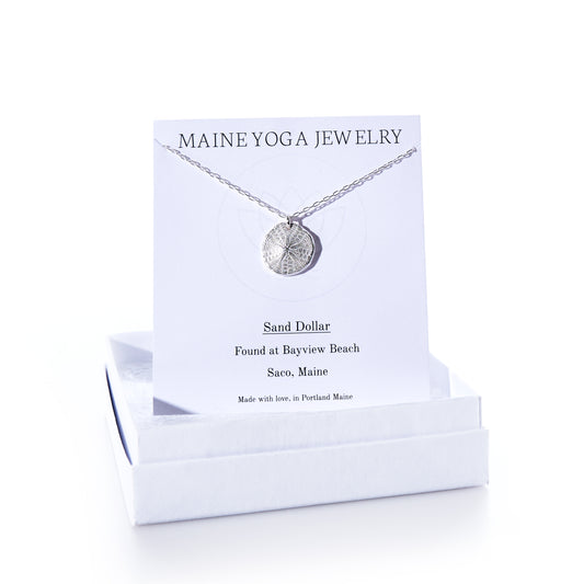 A Pendant Necklace - Sand Dollar Silver by Maine Yoga Jewelry rests on a card inside a white box. The card notes that the sand dollar was found at Bayview Beach, Saco, Maine, emphasizing the brands sustainable craftsmanship inspired by Maines natural beauty. by Sea Bags