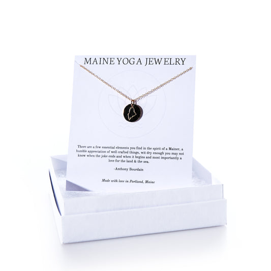 Pendant Necklace - Gold Maine by Maine Yoga Jewelry features a handcrafted round black pendant made from recycled sailcloth, elegantly suspended on a gold necklace. It comes displayed on a white card labeled “Maine Yoga Jewelry,” beautifully presented in a white jewelry box. Thoughtfully made in Maine, this sustainable piece combines eco-friendly materials with sophisticated design. by Sea Bags