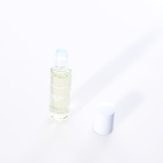 A clear glass roll-on bottle of Shore Soap Cos Warm Sand Eco-Friendly Roll-On Fragrance, featuring an alcohol-free jasmine scent, with a white plastic cap placed beside it on a white background. by Sea Bags