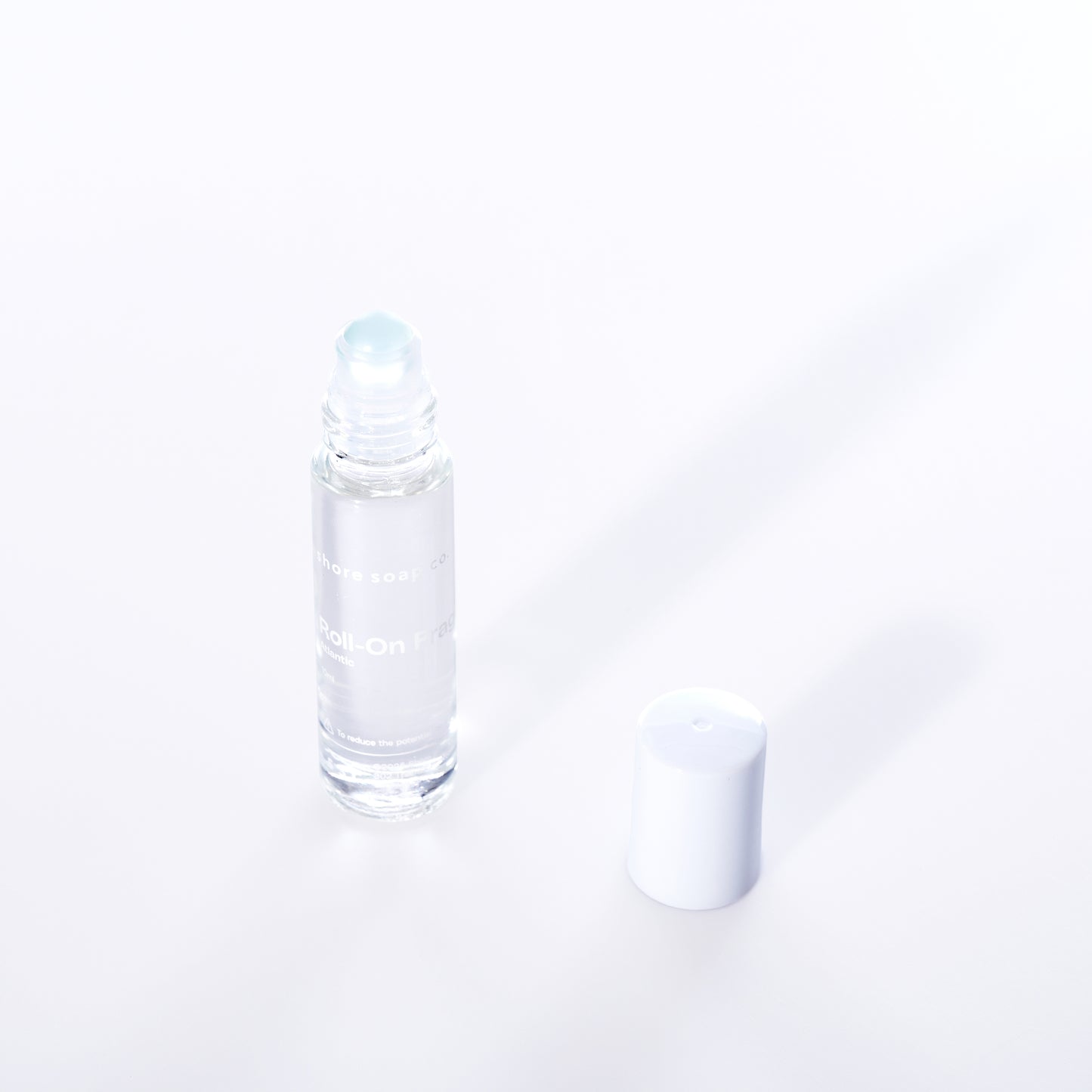 The Atlantic Eco-Friendly Roll-On Fragrance by Shore Soap Co is shown in a clear glass bottle with a white cap placed beside it on a white background, making it perfect for carrying your favorite alcohol-free perfume as a convenient travel fragrance. by Sea Bags