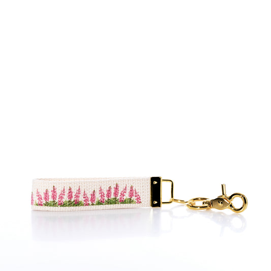 The Rustic Marlin Keychain - Pink Lupine by Rustic Marlin is a cream canvas keychain adorned with delicate pink lupine-inspired floral embroidery, gleaming gold hardware, and a lobster clasp—an elegant keepsake beautifully displayed on a white background with a soft reflection. by Sea Bags
