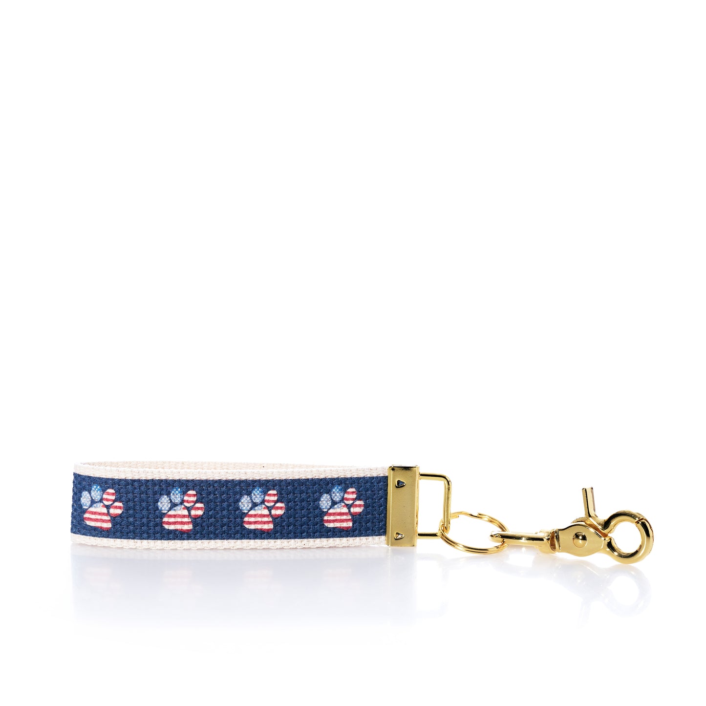 The Rustic Marlin Keychain - American Flag Paw from Rustic Marlin features a navy blue strip with American flag paw prints on a white background, complemented by a gold clasp and hardware—making it the perfect pawprint bag charm or dog keychain for any pet lover. by Sea Bags