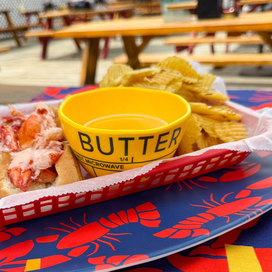 A Merritt Designs Butter Dipping Bowl in yellow sits between lobster meat on a bun and ridged potato chips in a red basket atop a lobster-themed tablecloth, capturing the essence of coastal dining with picnic tables visible in the background. by Sea Bags