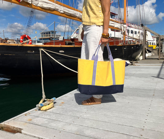 Person in white pants and sandals stands on a dock holding the Sea Bags Vintage Yellow Canvas Large Tote, with a sailboat moored in the background. by Sea Bags