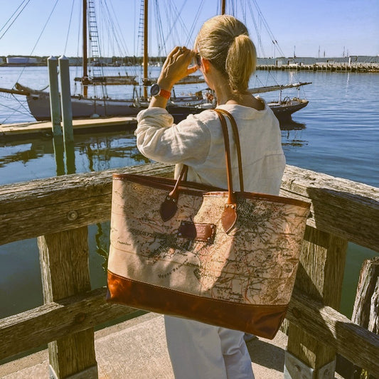 A woman carrying the Sea Bags Heritage Aello Large Tote stands on a dock, gazing at sailboats drifting on calm water beneath clear skies—including the beautifully restored S/Y AELLO, an iconic Greek sailing yacht celebrated for its classic heritage.