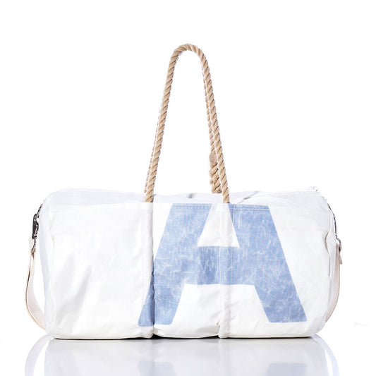 Introducing the Sea Bags Vintage Blue U Duffel—a white vintage duffel bag crafted from recycled sail cloth, featuring a bold blue letter U on the front. This stylish bag is finished with sturdy hemp rope handles and a beige adjustable shoulder strap, and is photographed against a crisp white background. by Sea Bags