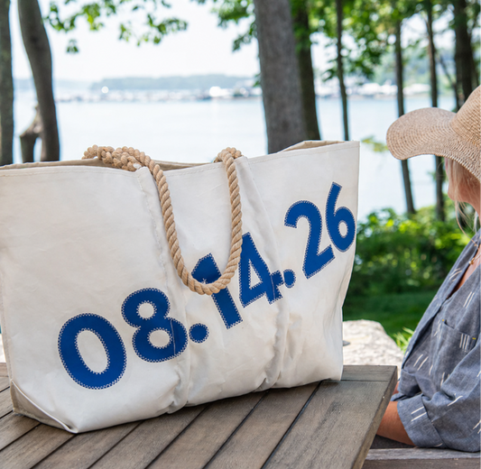 A large white Sea Bags Custom Date Tote featuring blue numbers 08.14.26 and rope handles rests on a wooden table outdoors near a hat-wearing individual, with trees and water visible in the background. by Sea Bags