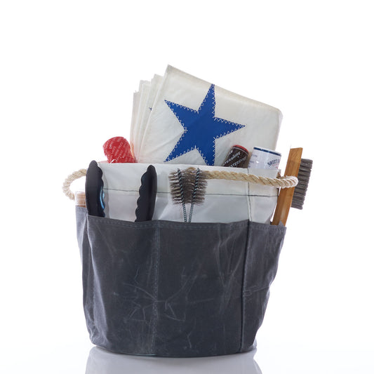 A Sea Bags All Purpose Gray Tool Bucket Bag holds a folded sail with a blue star, tongs, brushes, a bottle, and various barbecue and gardening tools against a white background. by Sea Bags