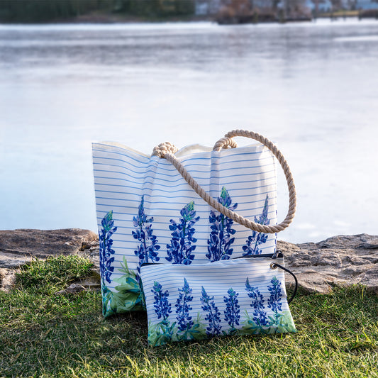 The Sea Bags Lupine Sailor Stripe Large Wristlet, crafted from recycled sail cloth and featuring blue floral and lupine print designs with coastal charm, is shown placed on grass near a lake with rocks and water in the background. by Sea Bags