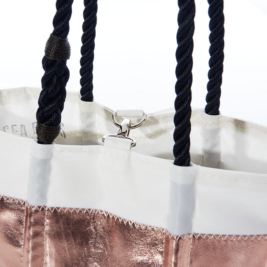 A close-up of the Sea Bags Rose Gold-on-Navy Mariner Stripe Handbag, crafted from durable sail cloth and featuring black rope handles, a metallic rose gold exterior with navy stripes, and white trim with a metal clasp inside. by Sea Bags