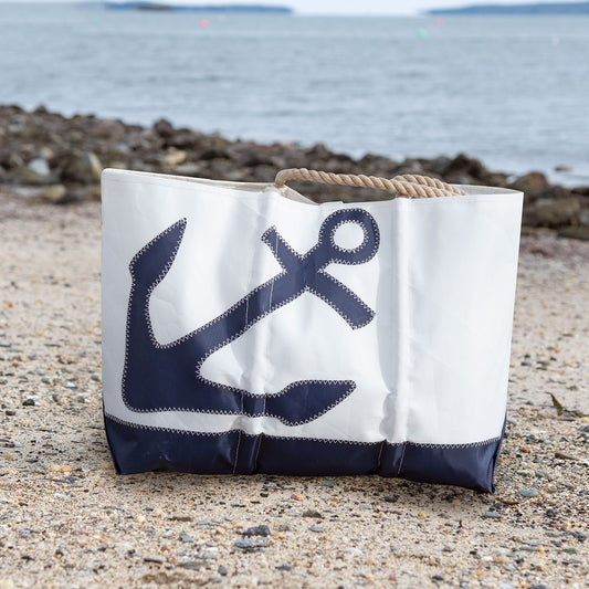 The Sea Bags Navy Anchor Ogunquit Beach Tote, made from recycled sail cloth and featuring rope handles and a classic navy anchor design, rests on a rocky beach near the water with distant land in view. by Sea Bags