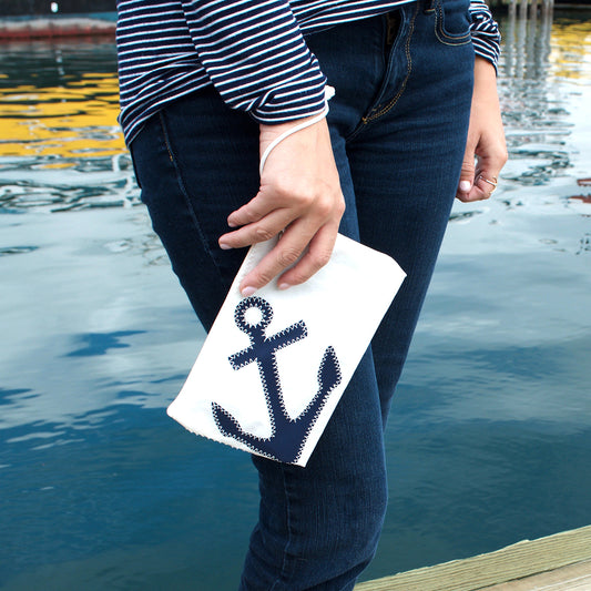 A person in a striped shirt and jeans holds the Sea Bags Navy Anchor Wristlet, crafted from recycled sail bags with a secure zip-top closure and featuring a navy blue anchor design, standing near water with reflections visible. by Sea Bags