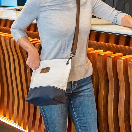 A person stands at a counter with a wavy wooden design, wearing blue jeans, a light gray top, and carrying the Sea Bags Chebeague Crossbody Bag—crafted from durable Dacron sailcloth and finished with a premium leather handle. by Sea Bags