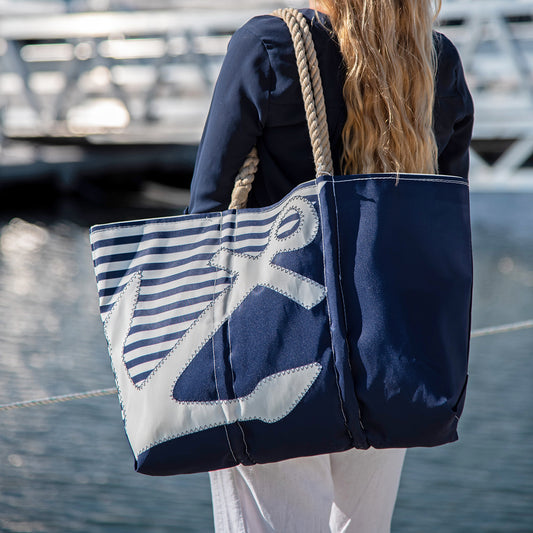 A person with long hair stands near the water, carrying the Sea Bags Breton Stripe White Anchor Large Tote—an eco-friendly navy-blue nautical bag with a white anchor design and rope handles, crafted from recycled sail cloth. by Sea Bags