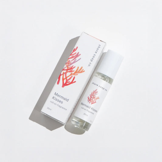 A 30ml Mermaid Kisses Eco-Friendly Roll-On Fragrance by Shore Soap Co is displayed alongside its packaging, which features an orange and pink coral illustration. This natural fragrance evokes the essence of ocean breezes in a convenient roll-on format. by Sea Bags