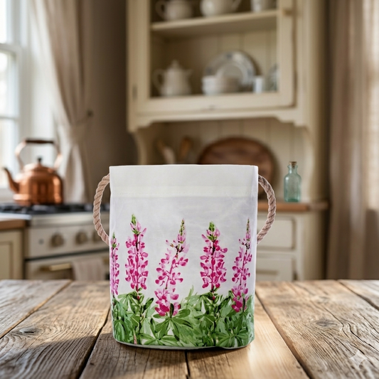 A Sea Bags Pink Lupine Bucket, featuring pink floral prints and rope handles, perfect for spring décor, sits on a wooden table in a rustic kitchen. by Sea Bags
