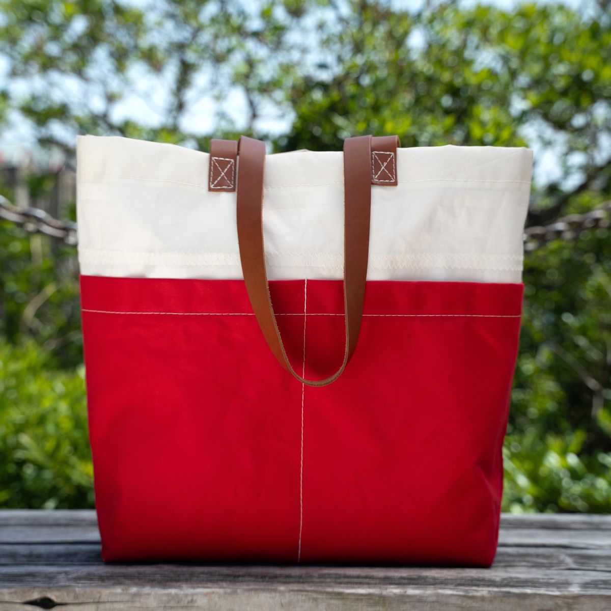 A Red Chebeague Market Tote by Sea Bags, crafted from recycled sails and finished with brown leather handles, rests on a wooden surface outdoors with greenery in the background. by Sea Bags