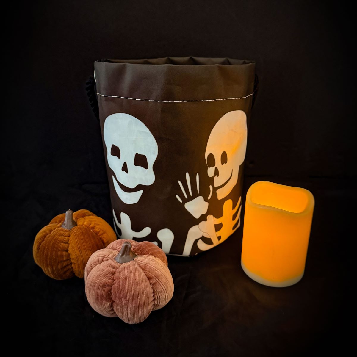 A Sea Bags Bucket of Bones Bag is displayed with skeleton graphics, accompanied by two plush pumpkins and a yellow LED candle, all arranged on a black surface against a dark background.