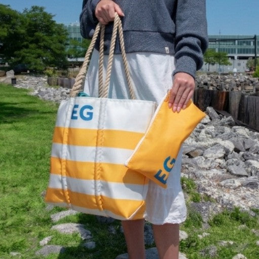 A person stands on the grass holding the Sea Bags Custom Monogram Breton Stripe Medium Tote Large Wristlet Bundle, crafted from recycled sailcloth and featuring a matching pouch; both items display an “EG” monogram. Rocks and trees in the background emphasize this personalized tote bag bundle’s unique charm.