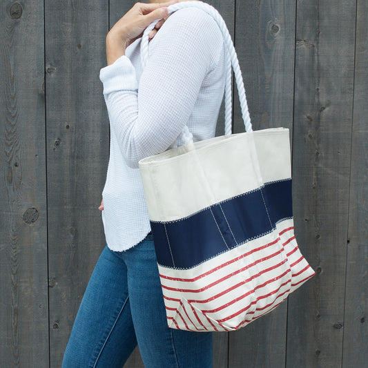 A person wearing a white long-sleeve shirt and blue jeans stands in front of a wooden fence, carrying the Sea Bags Red Mariner Stripe Medium Tote—a nautical-inspired bag crafted from recycled sailcloth in cream, navy, and red stripes—over their shoulder. by Sea Bags