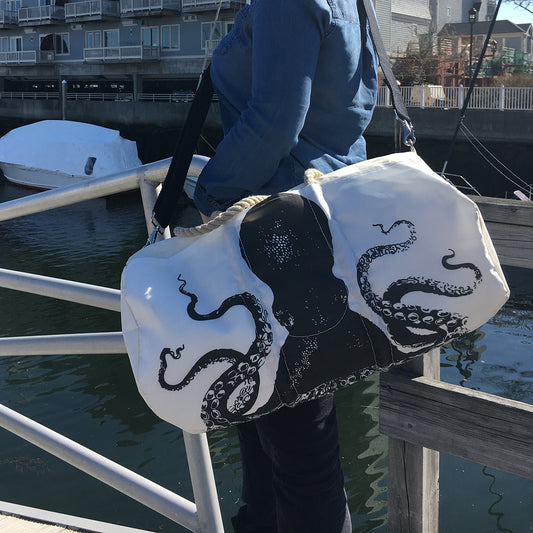 A person stands by a dock holding the Sea Bags Octopus Duffel, crafted from recycled sail cloth and featuring a marine grade zipper with a striking black octopus design, while boats and buildings can be seen in the background. by Sea Bags