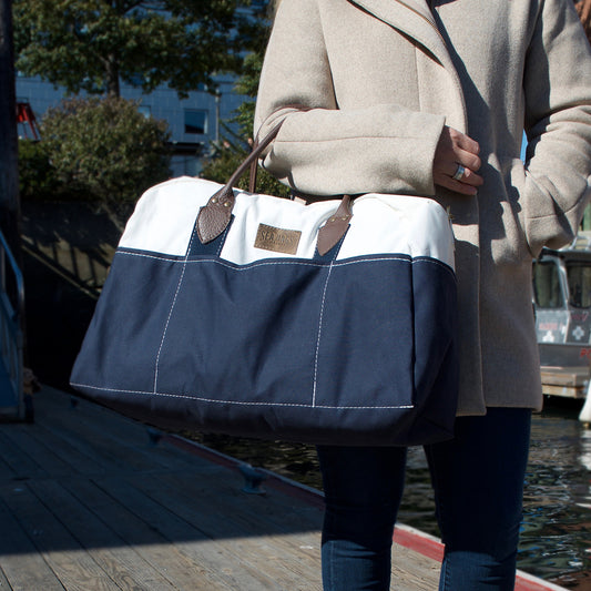 A person wearing a tan coat stands on a wooden dock near the water, holding the Sea Bags Chebeague Weekender—a spacious blue and white bag crafted from recycled sail cloth with brown leather handles. by Sea Bags