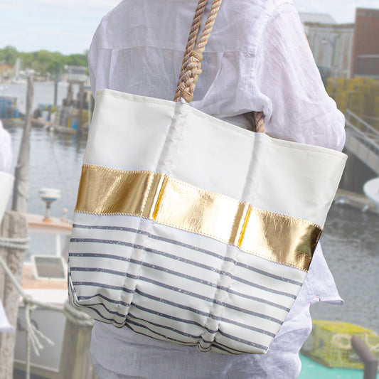 A person in a white shirt carries the Sea Bags Grey Mariner Stripe Tote—a metallic leather bag featuring gold and silver Breton-inspired stripes—while standing near a marina lined with boats and coastal buildings, perfectly complementing the nautical atmosphere. by Sea Bags