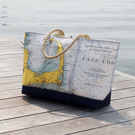 A Custom Design Ogunquit Beach Tote by Sea Bags, made from reclaimed sailcloth with rope handles and adorned with a nautical map of Cape Cod, rests on a wooden dock by the water—perfect as your personalized tote or Ogunquit Beach bag for your next seaside adventure. by Sea Bags