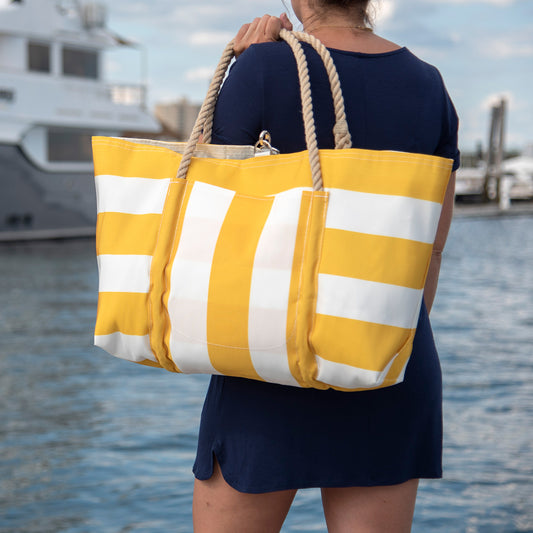 A person in a navy blue dress carries the Yellow Pier Tote by Sea Bags—a large yellow and white striped bag with rope handles and a spacious exterior pocket, crafted from recycled sail cloth—near a marina with boats in the background. by Sea Bags