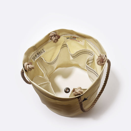 The Maine Bicentennial Beverage Bucket by Sea Bags is shown in top view, featuring an empty beige canvas design crafted from recycled sail cloth, rope handles, several inner pockets, and a visible metal grommet on the bottom. A SEA BAGS label is displayed inside. Inspired by Maine’s 1901 state flag heritage, this versatile bucket is ideal for storage or as a collapsible cooler. by Sea Bags