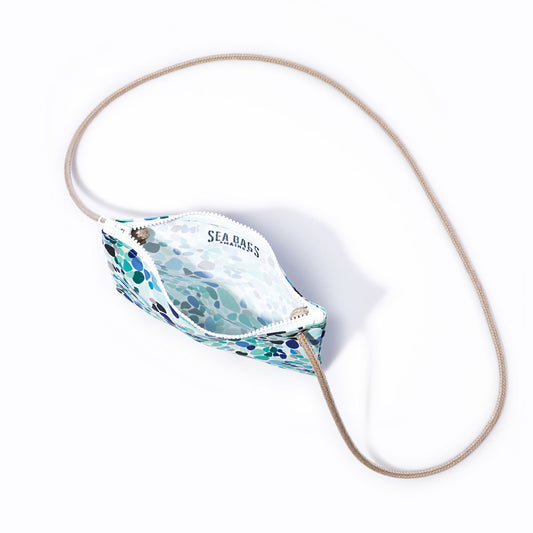 The Sea Bags Sea Glass Print Slim Crossbody is a compact pouch featuring a blue and green mosaic pattern inspired by sea glass tones. Crafted from recycled sail cloth, it includes a tan cord strap and an interior SEA BASS label. Shown on a white background, this accessory is one of our most striking designs. by Sea Bags