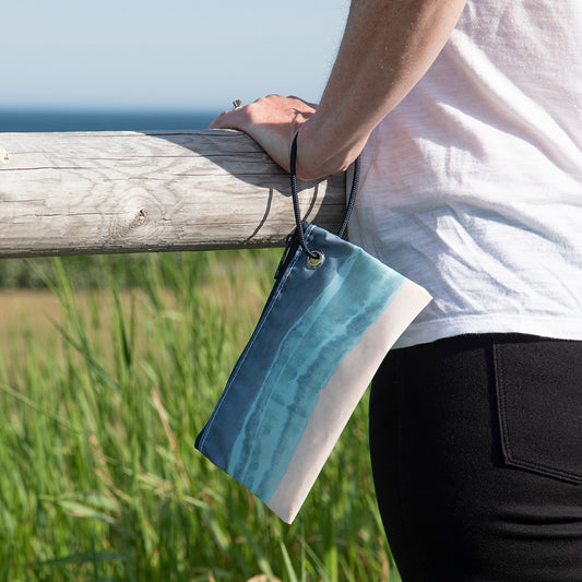 A person stands by a wooden fence holding the Sea Bags Shoreline Tie Dye Wristlet, a small zippered pouch crafted from recycled sails and featuring a blue ocean and sandy beach design with a convenient wrist strap. by Sea Bags