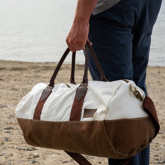 A person holding the Sea Bags Tan Chebeague Duffel, featuring white and brown details with leather straps, stands on a sandy beach near the water. by Sea Bags