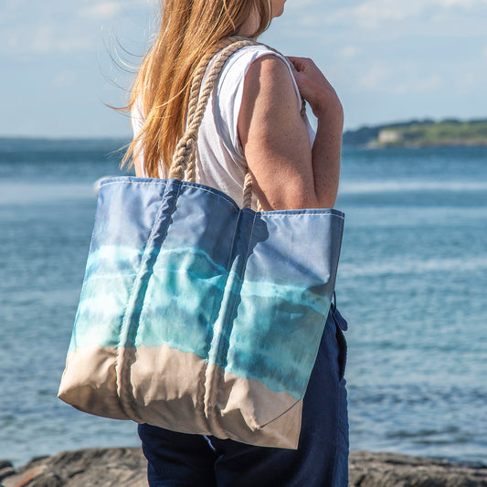 A person with long hair stands near a body of water on a sunny day, holding the Sea Bags Zip Top Shoreline Tie Dye Medium Tote, crafted from recycled sail cloth with rope handles in shades of blue and beige. by Sea Bags