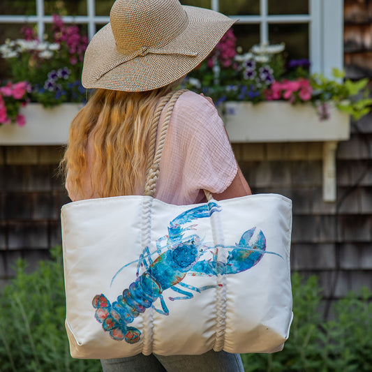 A person with long hair, dressed in a wide-brimmed hat and pink shirt, carries the Sea Bags Multicolor Lobster Large Tote, which features a blue cotton candy lobster design on white recycled sail cloth. Flowers and a window can be seen in the background. by Sea Bags