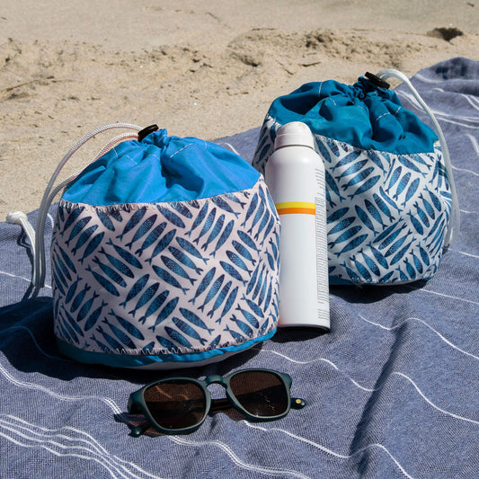 Two Sea Bags Fish Print Ditty Bags, including a weather-resistant style, sit on a towel in the sand next to a bottle of sunscreen and a pair of sunglasses. by Sea Bags