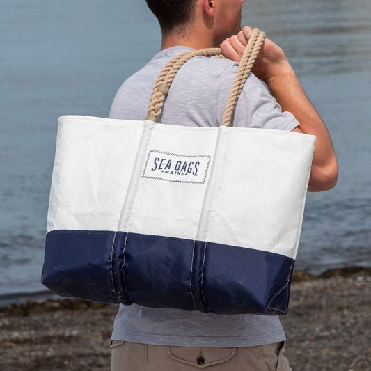 A person holds the Sea Bags Sailmakers Tote with Clasp and Hanging Pocket, a large white and navy blue nautical bag with rope handles made from recycled sail cloth, standing near water. by Sea Bags