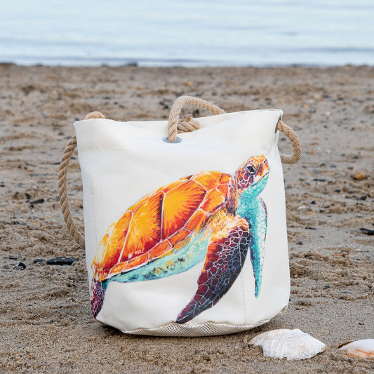 The Multicolor Sea Turtle Beachcomber Bucket by Sea Bags, crafted from recycled sails and featuring a vibrant sea turtle design, rests on the sandy beach near seashells with the ocean in the background. by Sea Bags
