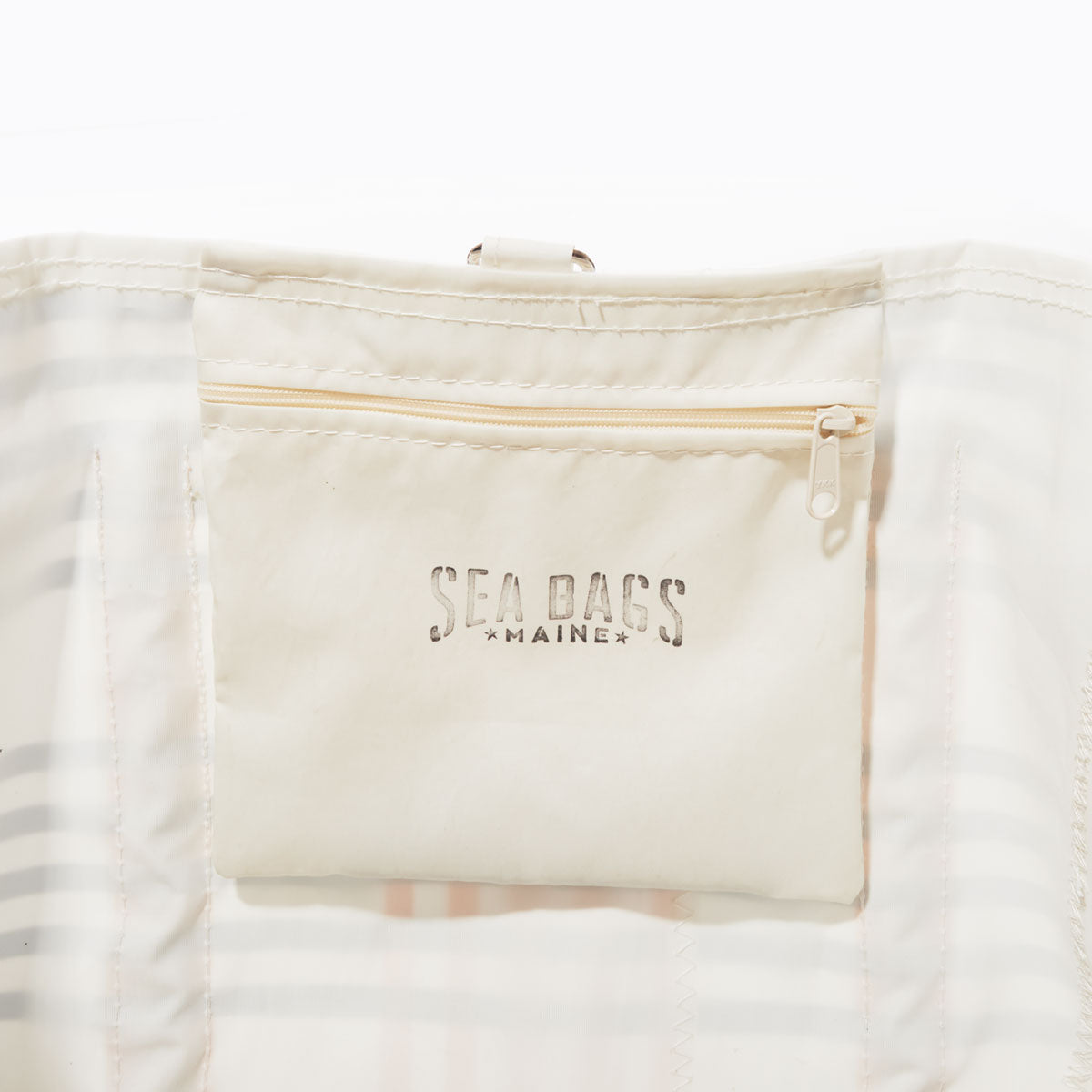 Close-up of a cream-colored zippered pouch with Sea Bags printed on it, attached to the interior of the weatherproof Picnic Stripe Pier Tote, which features a light plaid lining. by Sea Bags