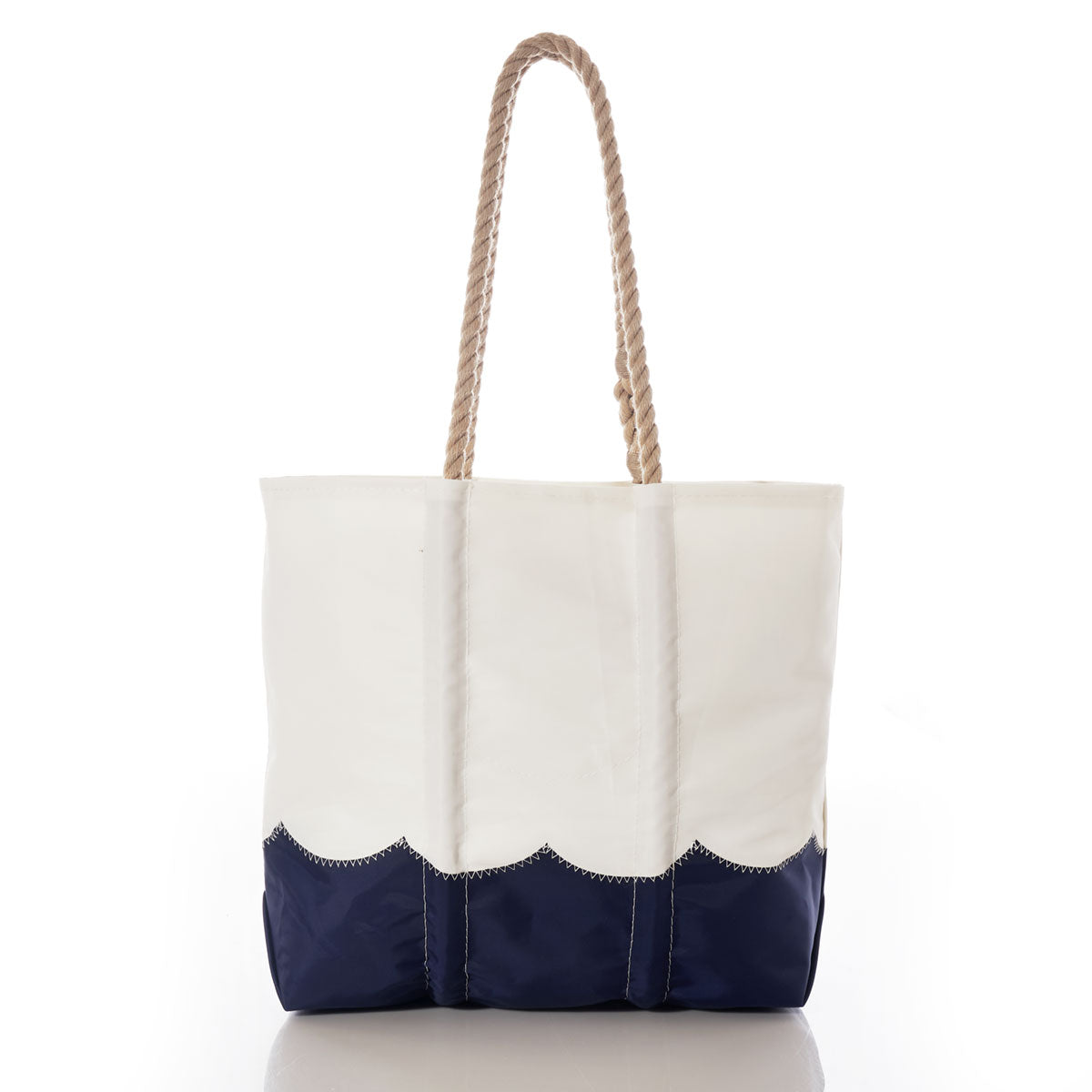Sea Bags Wavy Seas Medium Tote—a white tote bag featuring a navy blue scalloped bottom, visible stitching, and two sturdy rope handles, crafted from recycled sail cloth for long-lasting durability. This classic nautical beach bag is shown against a white background. by Sea Bags