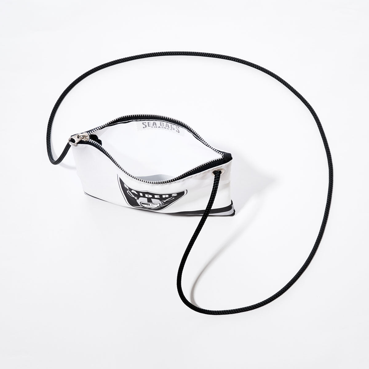 The Las Vegas Raiders Slim Crossbody by Sea Bags is a small white bag made from recycled sail cloth, featuring a black cord strap and a distinctive black-and-white patch on the front inspired by the iconic NFL team. Presented against a plain white background, this unique crossbody blends sporty Raiders style with an eco-friendly design. by Sea Bags