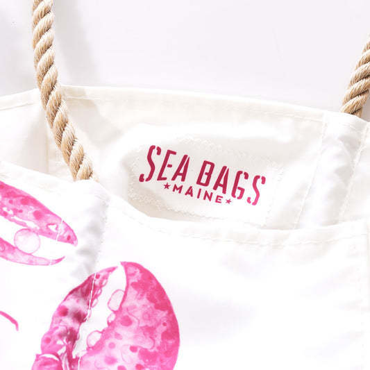 Close-up of the Sea Bags Pink Lobster Medium Tote, a white bag with sturdy rope handles, featuring a pink lobster print in a watercolor style by a Maine artist, and finished with the Sea Bags label. by Sea Bags