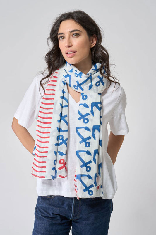A woman stands against a plain light gray background wearing a white shirt, jeans, and the Anchor Pop Scarf by Batela USA Inc—a nautical cotton scarf adorned with blue and red scissors and stripe patterns. by Sea Bags