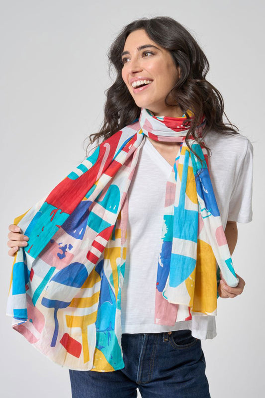 A woman wearing a white t-shirt and blue jeans smiles as she displays the Batela USA Inc Downeast Buoys Scarf—a vibrant, abstract-patterned cotton scarf—draped around her neck. by Sea Bags