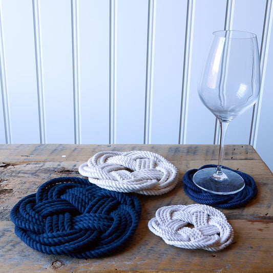 Four Nautical Sailor Knot Coasters-Navy by Mystic Knotwork, crafted in classic Turk’s head knot style with navy blue and white woven rope, are arranged on a wooden table, with a clear empty wine glass resting atop one of the coasters. These coasters lend maritime charm to your décor, set against a white paneled wall backdrop.