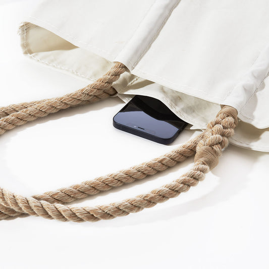 A black smartphone rests inside an open Sea Bags Acadia National Park Tote, crafted from white recycled sail cloth with thick rope handles, set against a white background and echoing the timeless style of coastal Maine. by Sea Bags