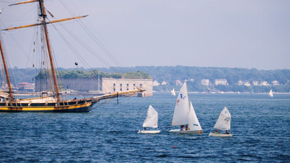 a sailing fleet in a coastal harbor flanked by a large sailing ship at anchor