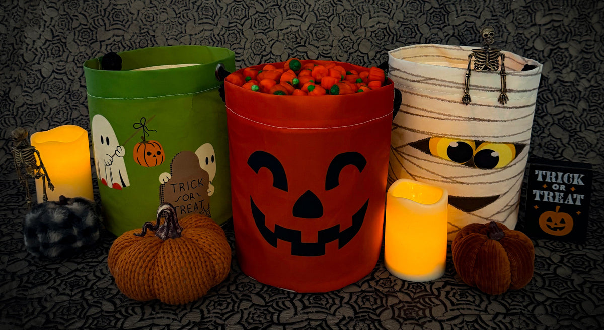 Three Halloween-themed buckets, a variety of pumpkins, two candles, and a “Trick or Treat” sign are arranged on a dark lace background. The center bucket is filled with candy pumpkins.